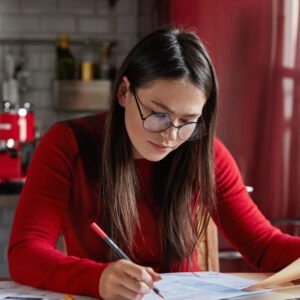 Busy female enterpriser thinks about target or planning company, makes statistics or analytic research, sits at desktop, kitchen interior in background, concentrated on work, wears round spectacles
